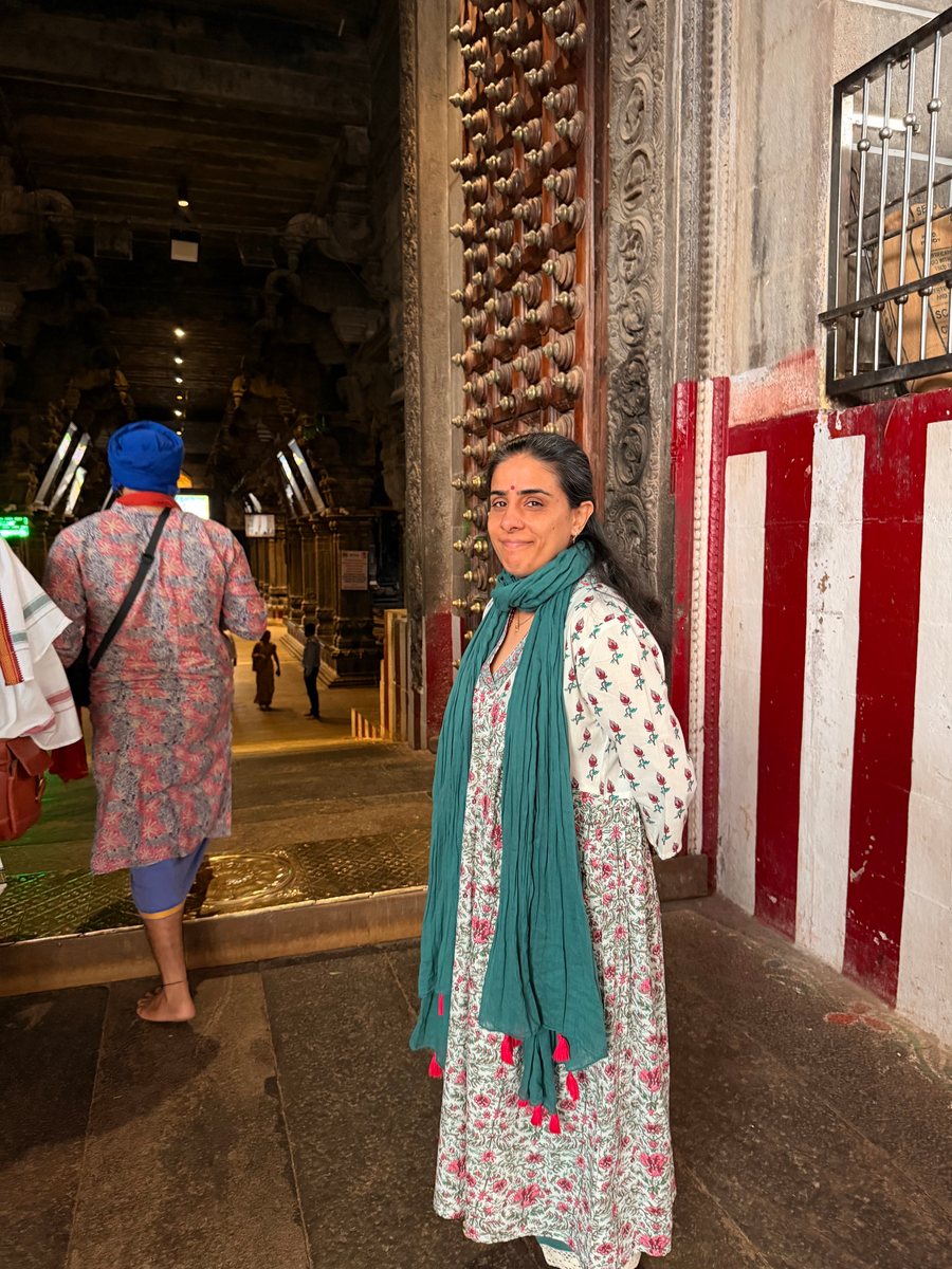 Kanika standing at the carved wooden doorway of a Tamil Nadu temple, smiling over her shoulder, the pillared corridor stretching into shadow behind her