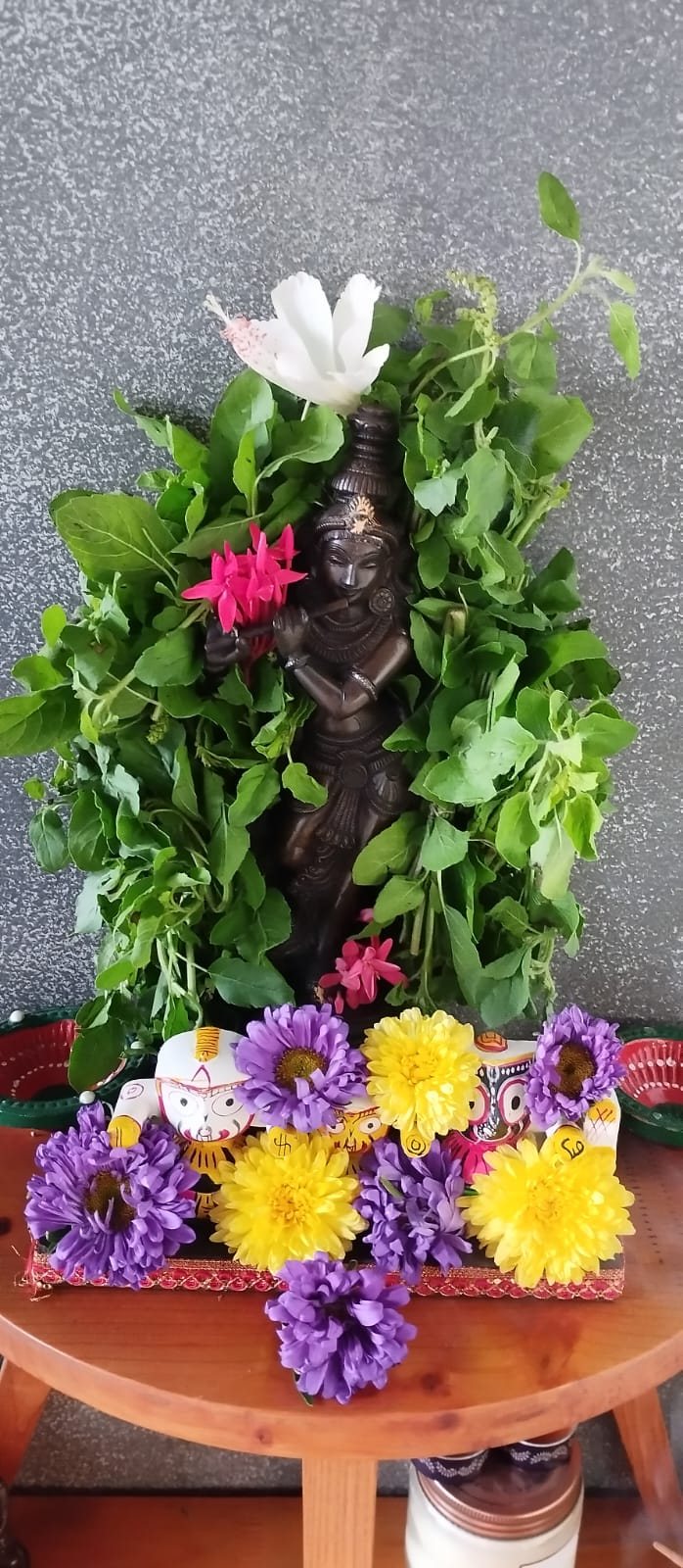 Krishna garlanded in tulsi with a white hibiscus above his crown and pink ixora at his shoulder, purple and yellow chrysanthemums banked along the painted Jagannath figures at his feet
