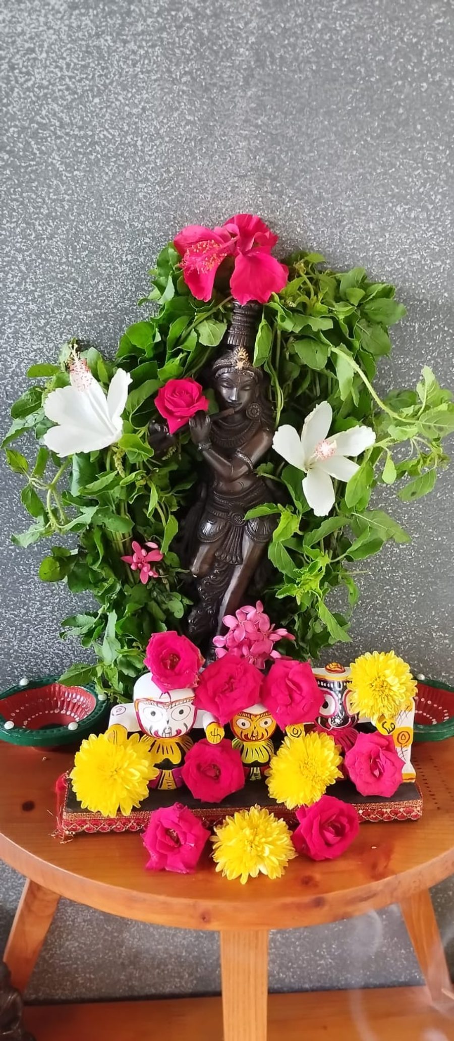 A dark brass Krishna playing the flute, garlanded in fresh tulsi leaves with pink roses, white hibiscus and yellow chrysanthemums, small painted Jagannath figures at his feet