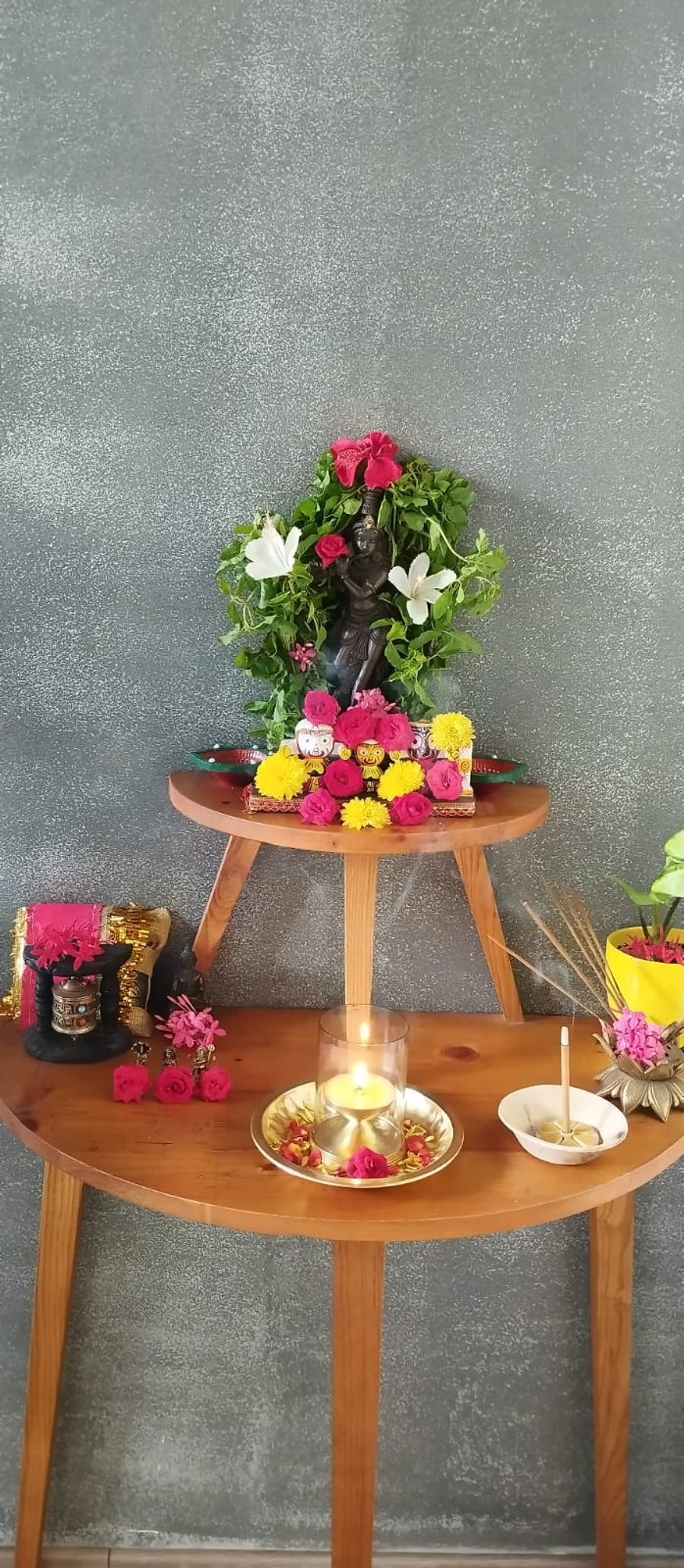 A garlanded brass Krishna on a small wooden table against a grey wall, with a lit lamp in a glass, incense smoke rising, flowers and a money plant around