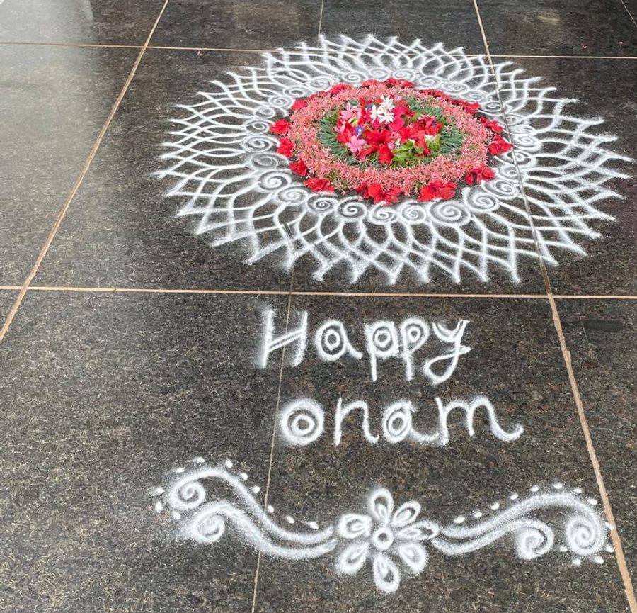 A large circular kolam drawn in white rice-flour lace on dark granite, its centre filled with a pookalam of red, pink and white flowers, with 'Happy Onam' written below in white powder