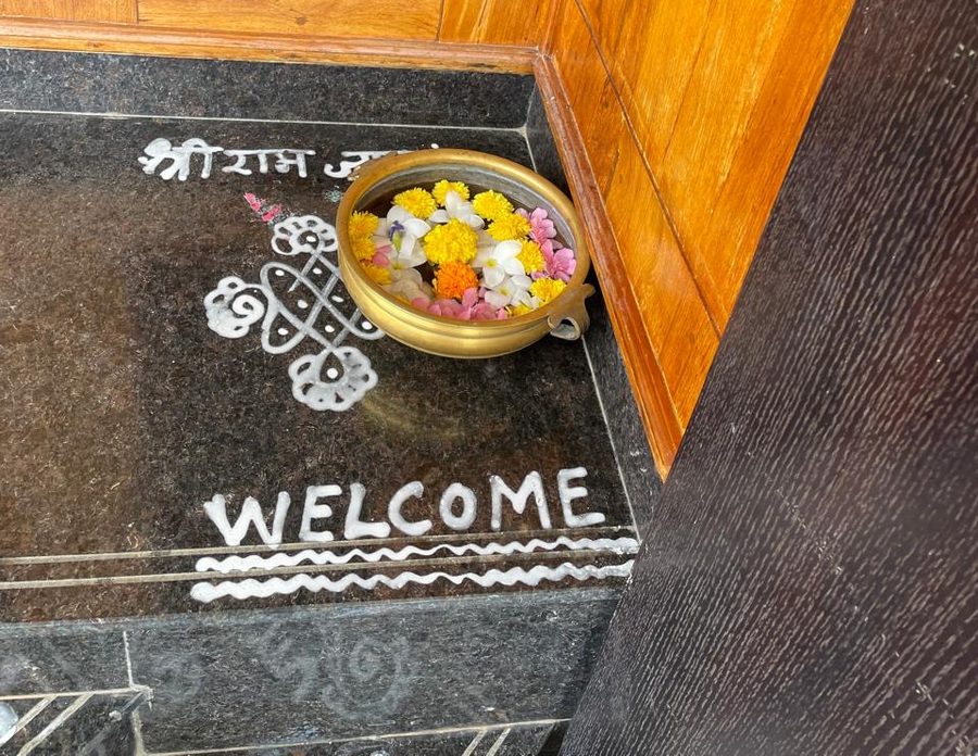 A brass uruli filled with marigolds, jasmine and pink blossoms floating on water, set on a dark granite doorstep beside a kolam and the word WELCOME