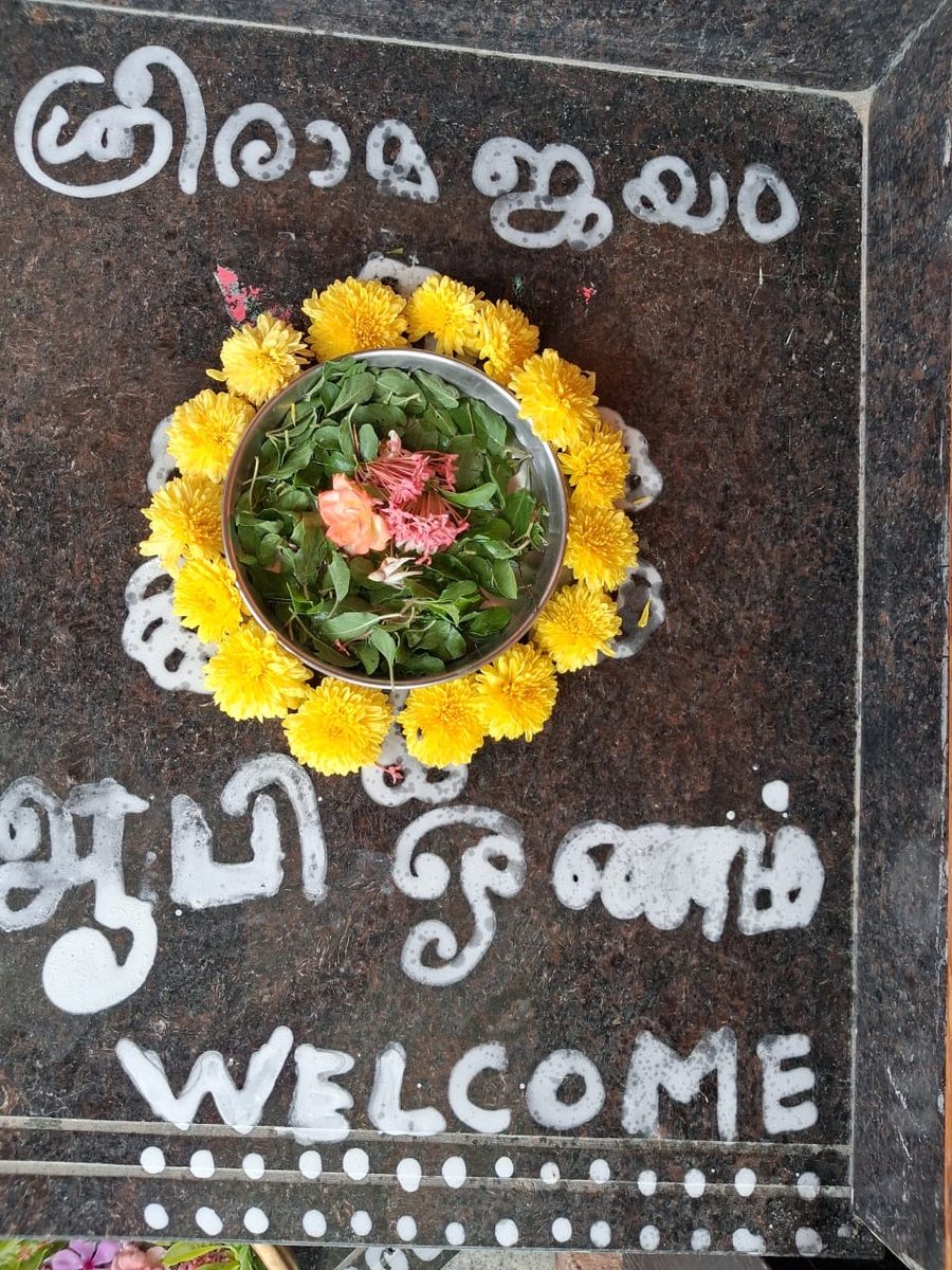 A doorstep decorated in white paste lettering — Malayalam script above, Tamil script and the word WELCOME below — around a steel bowl of curry leaves ringed with yellow chrysanthemums